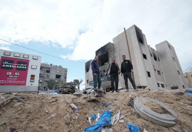 Three men stand surrounded by rubble and an ambulance.