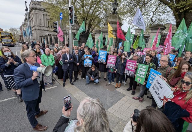 Crowd gathered outside Dáil Éireann with placards while man holds megaphone wearing a suit.