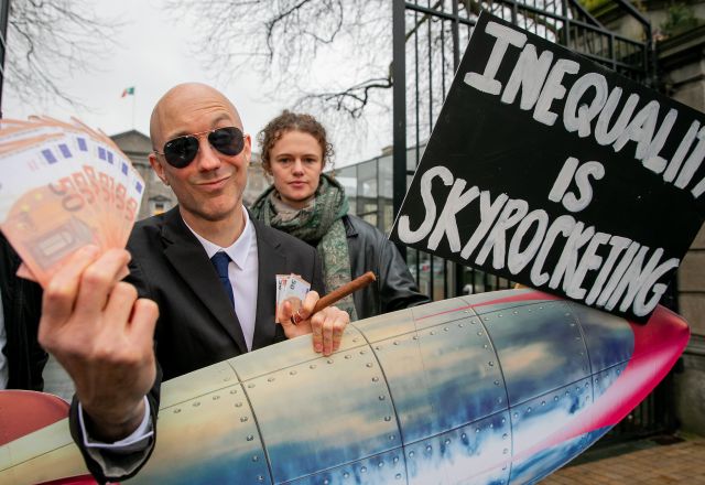 A man with a rocket, money and a cigar and a protester behind him with a sign saying inequality is skyrocketing