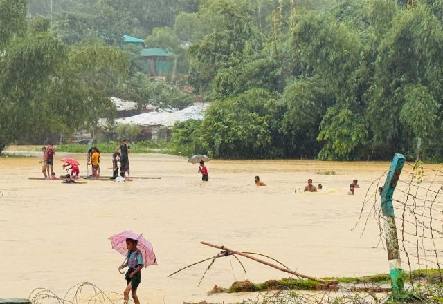People move through floodwater in Cox’s Bazar after heavy rain, with umbrellas visible near flooded shelters and trees.