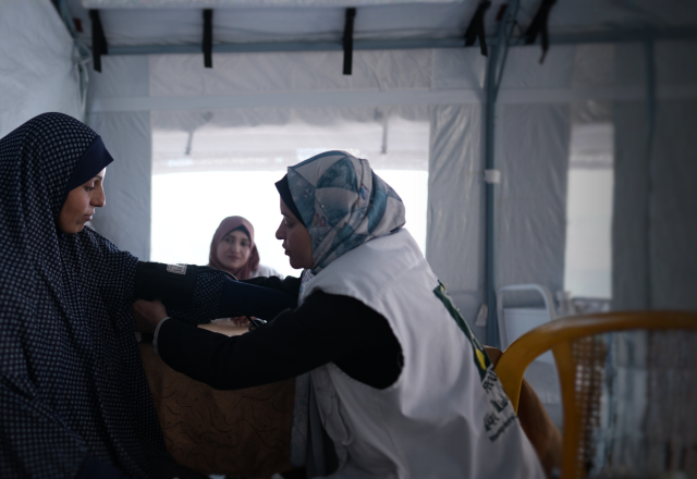 Three women are sitting in a white room. One woman, a doctor, is taking the blood pressure of another woman in the foreground