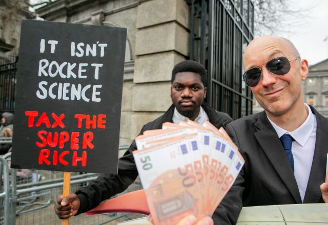 Two men hold money and a sign that says Tax The Super Rich