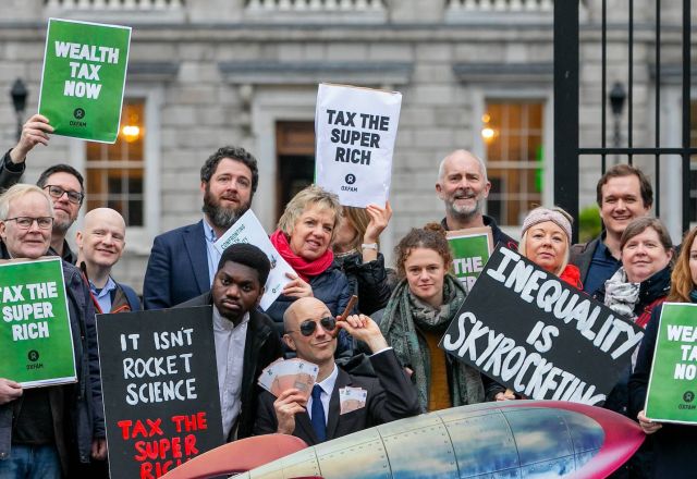 A group of people stand outside Leinster house holding signs that say Wealth Tax Now