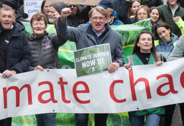 A group of people walk towards the camera carrying a white banner with red letters, saying climate chaos. There are green banners in the background