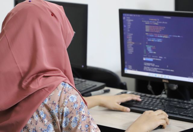 A lady in a pink headscarf works on a computer. Her hands are placed on the keyboard, the screen of the computer is blue