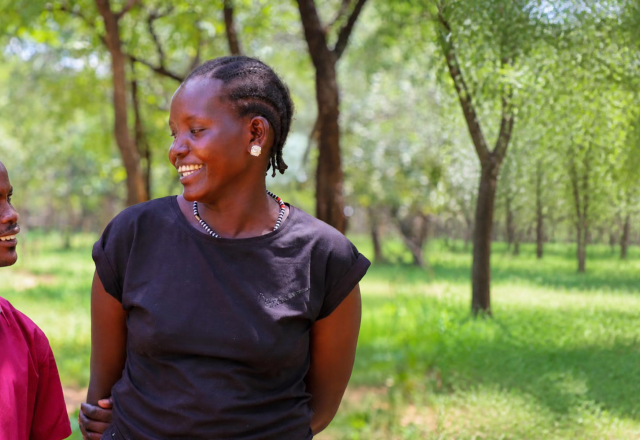 Two people stand in a wooded area, looking and smiling at each other