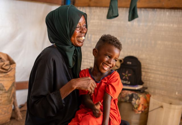 A lady and her child laugh. The lady wears a black hijab, her child wears a red shirt. They are in a tent