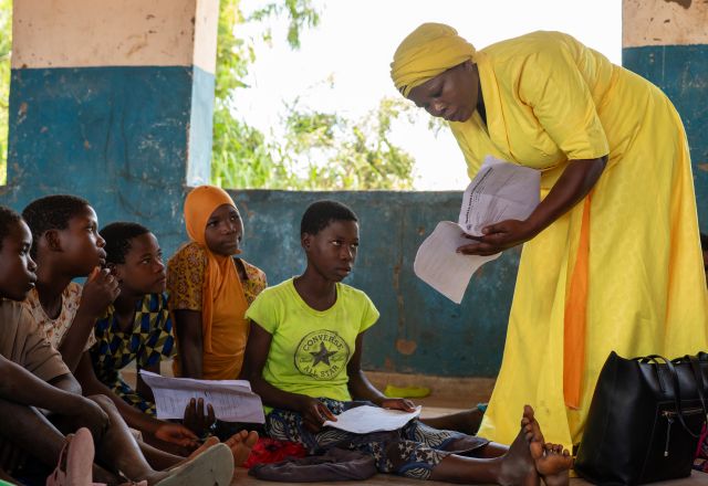 A lady dressed in yellow leans over to show children something on sheets of paper she is holding