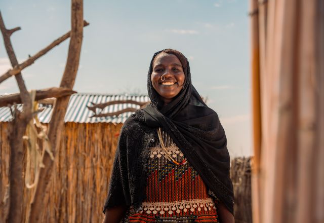 A lady smiles at the camera. She is wearing a black headscarf and colourful dress. There are stick fences around her