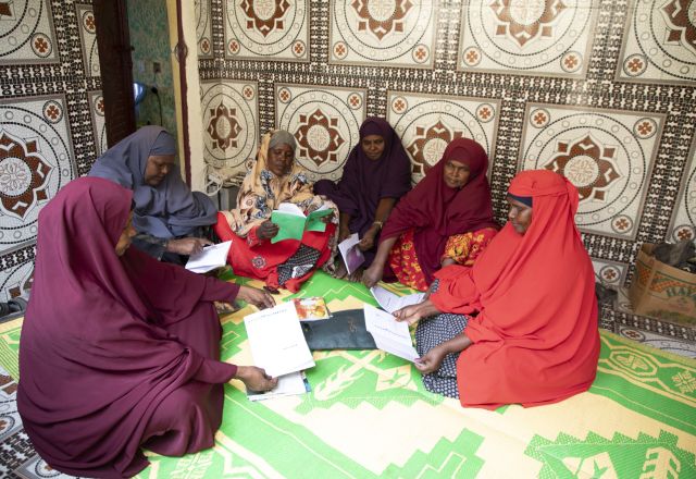 Women sit on the ground looking at books together. They are wearing different coloured hijabs