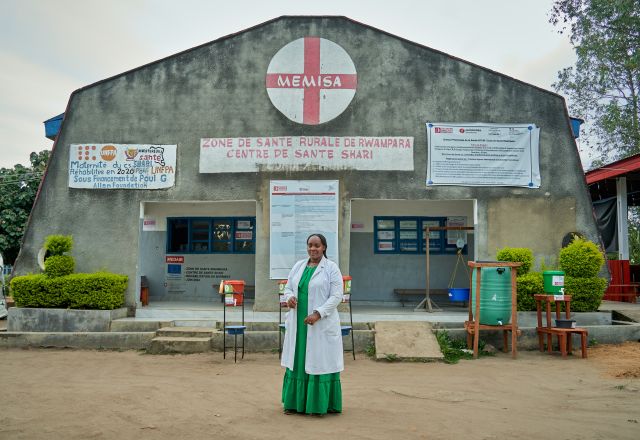 A lady in a green dress and long white coat stands in front of a hospital