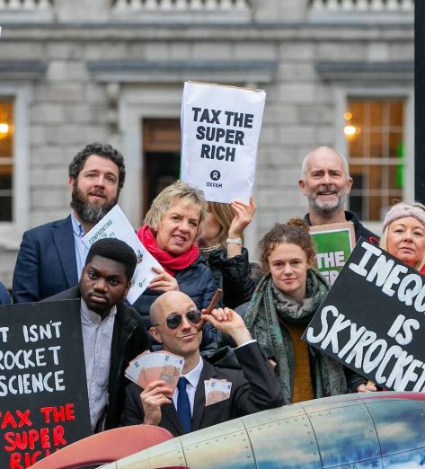 A group of people stand outside Leinster house holding signs that say Wealth Tax Now