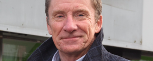A middle aged man wearing a dark coat with a high collar smiles into the camera.