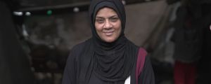 A lady in a black hijab smiles at the camera. She is in a makeshift tent camp in Gaza