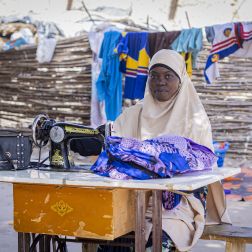 Zahara Seybou Harouna sitting outside, posing while working on sewing clothes with her sewing machine.