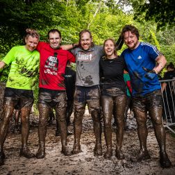 A group of 4 men and a women at the end of the Hell and Back Challenge. They are holding each other side by side, smiling and covered in mud.