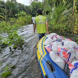 Oxfam staff pull a supply-laden inflatable boat through a flooded area surrounded by greenery.