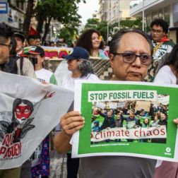 A man holding a sign reading “Stop Fossil Fuels – climate chaos” while standing in a crowded street protest with others carrying banners and flags.