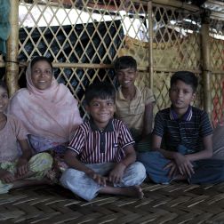 Romida sits with four children inside a bamboo shelter in Cox’s Bazar camp, where the family has lived for seven years after fleeing violence and faces safety concerns in poorly lit areas.
