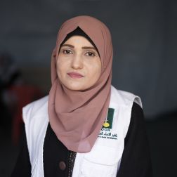 A female doctor looks into the camera. She is wearing a pink headscarf and wears a white vest over a dark top