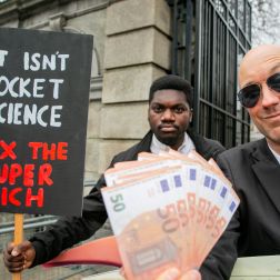 Two men hold money and a sign that says Tax The Super Rich