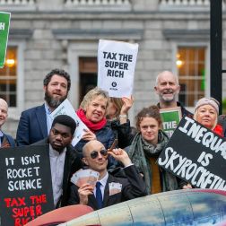 A group of people stand outside Leinster house holding signs that say Wealth Tax Now