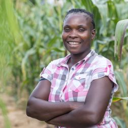 A lady stands in a field wearing a short sleeved shirt. She is smiling at the camera