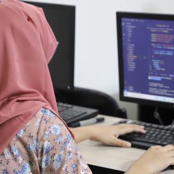 A lady in a pink headscarf works on a computer. Her hands are placed on the keyboard, the screen of the computer is blue