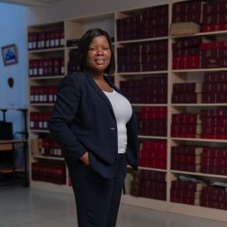 A lady wearing a dark business suit smiles at the camera. In the background is shelves with files and books
