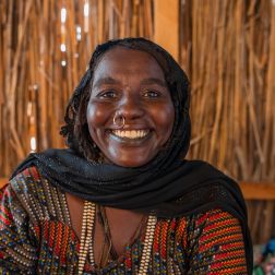 A lady smiles into the camera. She wears a coloured dress and black scarf