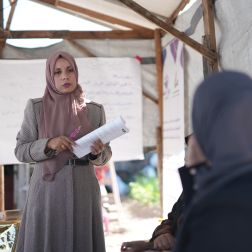 A lady in a pink headscarf holds a rolled up sheet of paper. Another person in dark clothes watches her, with their back to the camera