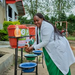 A lady washes her hands with water from an orange bucket. She is wearing a green dress and white overcoat