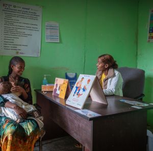 A young mother with her baby speaks to a smiling nurse in a green-walled clinic in Ethiopia, with health posters and educational materials visible.
