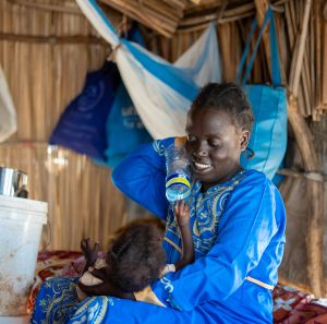 Ayul Ajak, a visually impaired Sudanese refugee mother, breastfeeds her baby inside a makeshift shelter with an Oxfam bucket nearby.